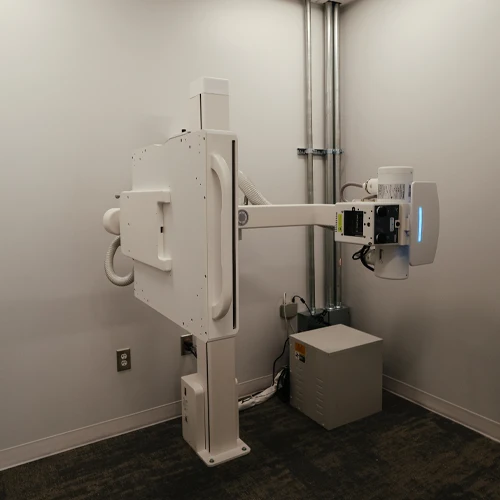 X-ray imaging machine positioned against a wall in a clinical room with visible utility connections.