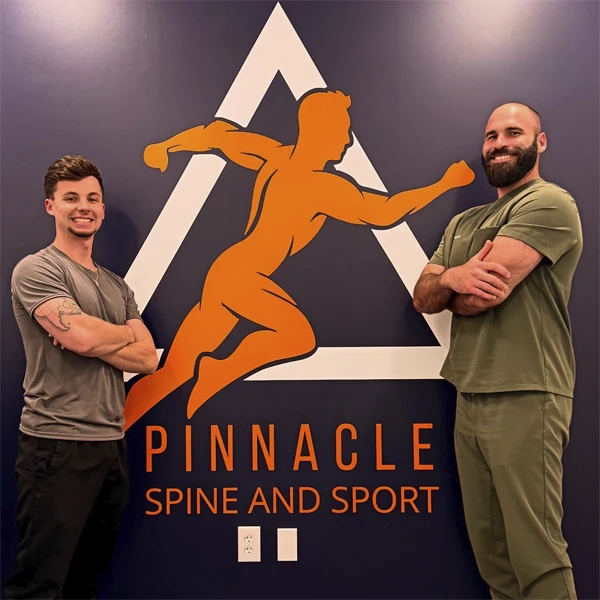 Two clinic team members standing in front of a wall featuring the Pinnacle Spine and Sport logo.